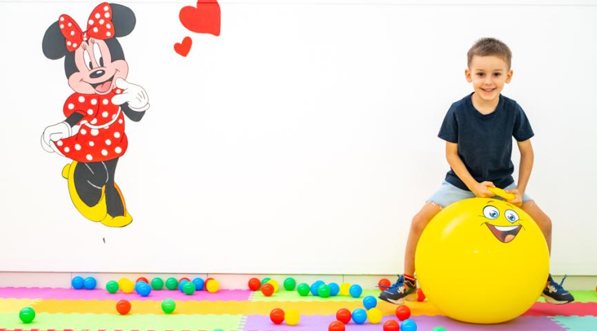 Kind spielt auf gelbem Ball, Minnie an der Wand.